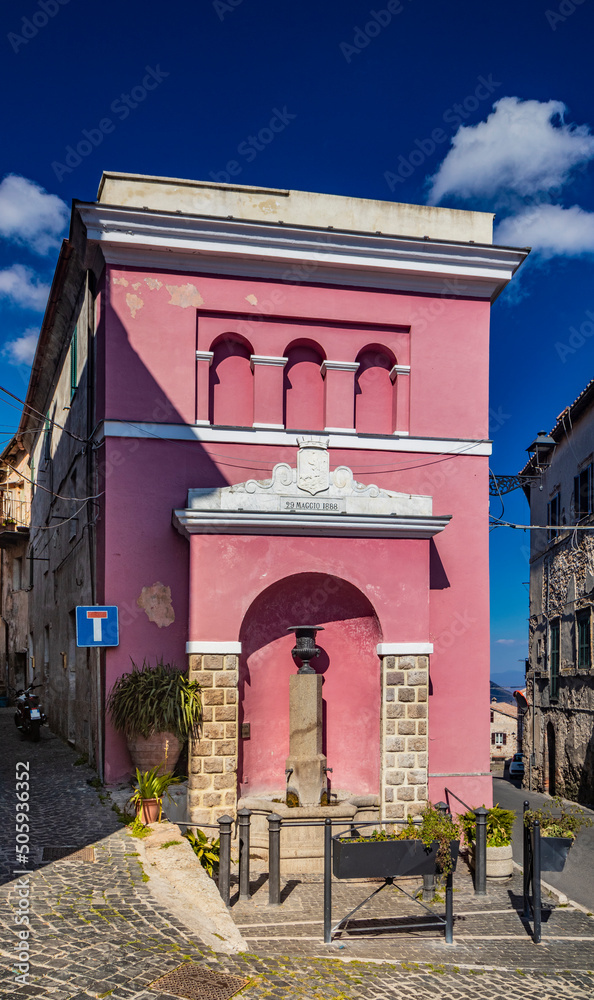 Fototapeta premium The small village of Tolfa, in Lazio. A glimpse of the town with its old stone and brick houses and cobbled alleys. An ancient fountain rests on the pink wall of a building.