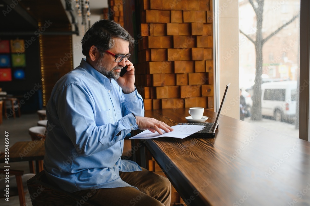 Male freelancer is working in a cafe on a new business project. Sits at a large window at the table. Looks at a laptop screen with a cup of coffee Male freelancer is working in a cafe on a new business project. Sits at a large window at the table. Looks at a laptop screen with a cup of coffee