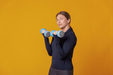 Workout concept. Young athletic woman in sportswear trains biceps muscles with dumbbells in her hands on a yellow background. Bodybuilding and fitness. Healthy lifestyle