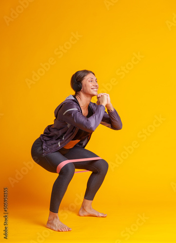 Workout concept. Athletic strong woman in sportswear practicing deep squat barefoot with fitness band on yellow background. Healthy lifestyle. Bodybuilding and fitness.