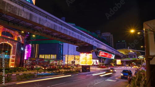 Timelapse of modern city intersection at night. Car light trails, bright urban lights, ads, people cross streets, monorail train station, Kuala Lumpur