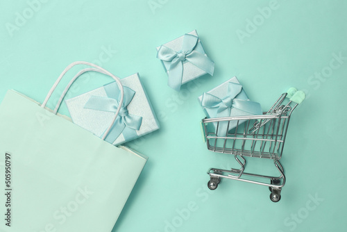 Paper shopping bag with gift boxes and shopping trilley on blue pastel background. Top view