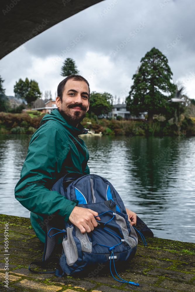 Young, bearded, happy and handsome guy with jacket and backpack and panoramic view of Calle-calle river, bridge, forest, and cloudy sky, Valdivia, Chile