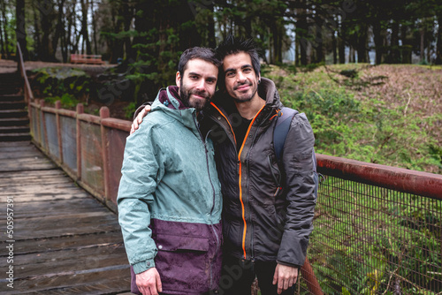 Two happy, young, caucasian, bearded and handsome brothers with jacket, cap and backpacks in a wooden bridge in the forest in a rainy day, Valdivia, Chile photo