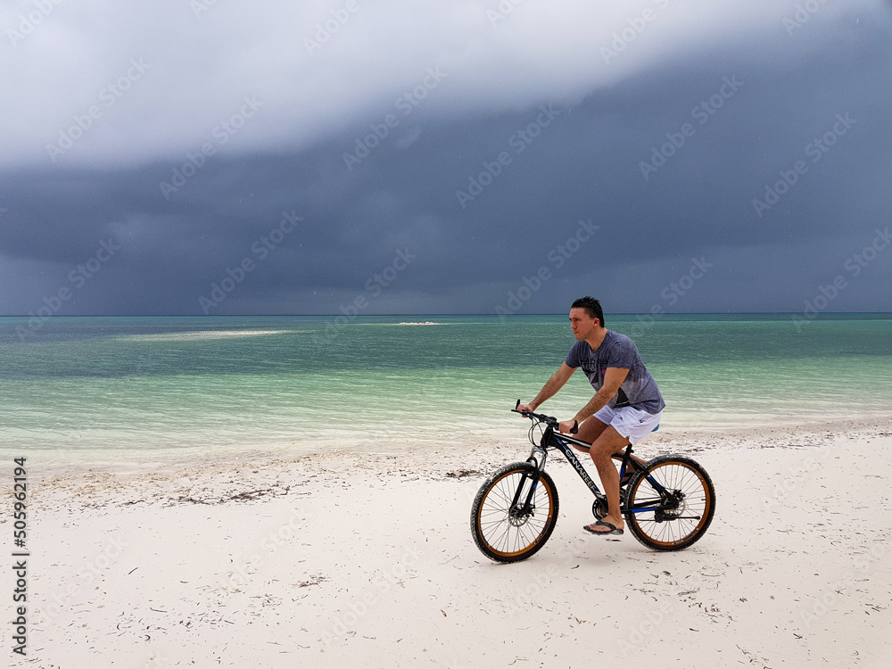man riding a bike on sand on tropical beach in stormy weather, fitness everywhere 