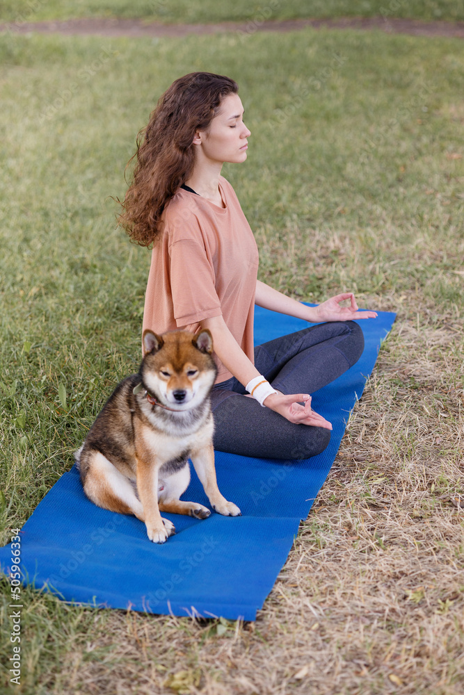 A European woman with curly hair meditates in the park with her dog on a blue yoga mat. meditation for mental and physical health. a woman sits in the lotus position and namaste with her eyes closed A European woman with curly hair meditates in the park with her dog on a blue yoga mat. meditation for mental and physical health. a woman sits in the lotus position and namaste with her eyes closed