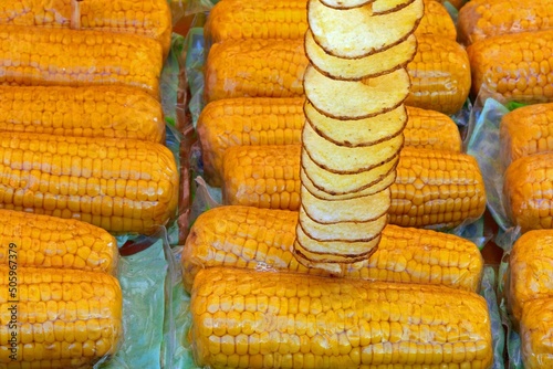 yellow corn cobs with plastic wrap and a skewer of fried potato chips with peel for sale in street food stall