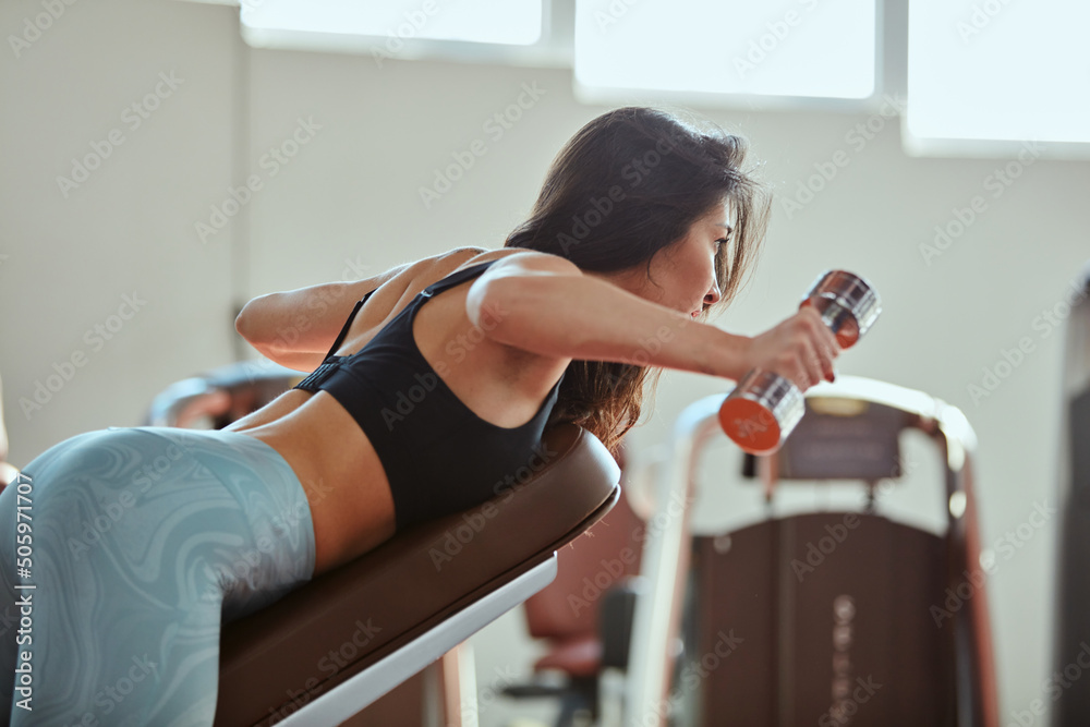 A fit, strong female bodybuilder is lifting dumbbells and doing exercises for biceps in a gym. - obrazy, fototapety, plakaty 