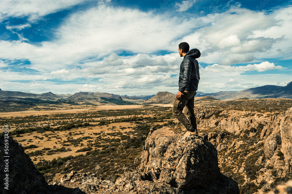 Young man standing on a rock at the top of a cliff overlooking a valley with a mountain range in the background. Concept of overcoming. Argentinian Patagonia