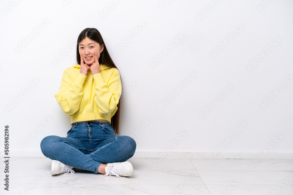 Young Chinese woman sitting on the floor isolated on white wall smiling with a happy and pleasant expression