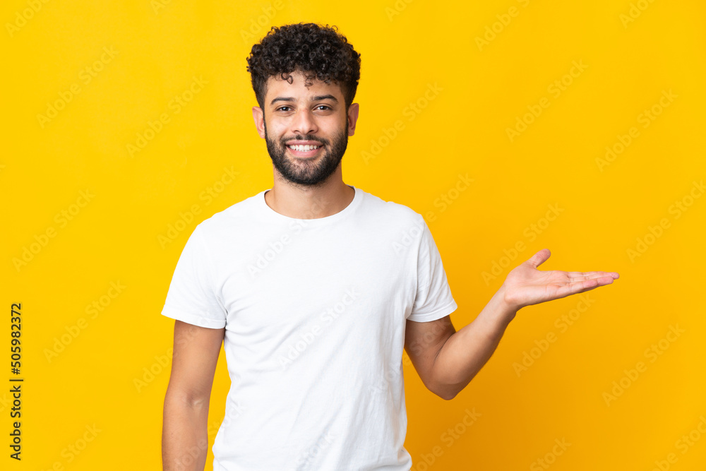 Young Moroccan man isolated on yellow background holding copyspace imaginary on the palm to insert an ad