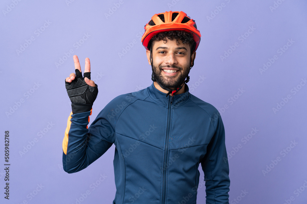Young Moroccan cyclist man isolated on purple background smiling and showing victory sign - obrazy, fototapety, plakaty 