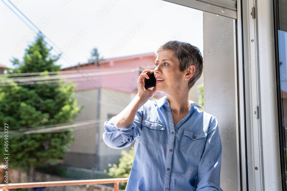 One woman mature caucasian female using mobile phone by the window at home in bright day or morning daily routine happy smile joyful and confident talking making a call copy space