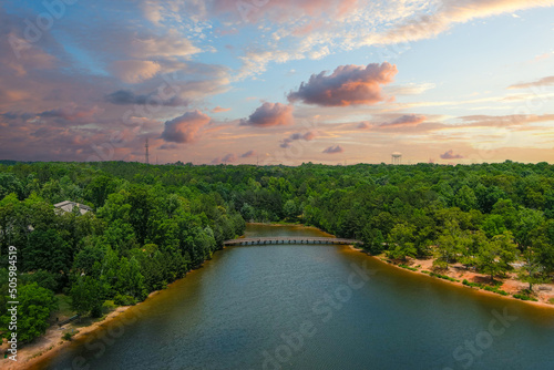 a gorgeous aerial shot of the rippling lake water surrounded by lush green trees, grass and plants with a gorgeous powerful clouds at sunset and a brown wooden bridge over the water at Indian Lake photo