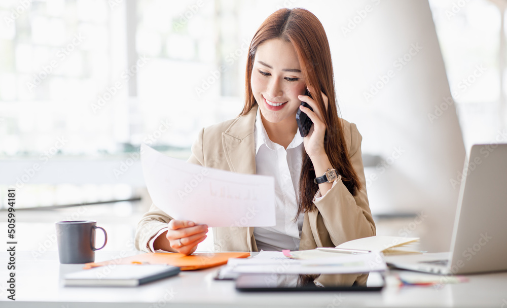 Asian businesswomen have the joy of talking on the phone with a laptop computer tablet on the office desk. doing accountant on a calculator to calculate business data documents.