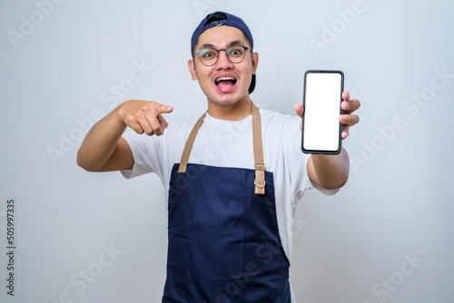 Young handsome asian barista man wearing apron pointing at mobile screen, showing app and smilling