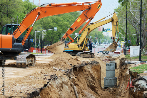 Reconstruction of the underground sewerage system on a city street in spring. Two excavators are digging a deep ditch. Traffic has been halted on a section of the street.