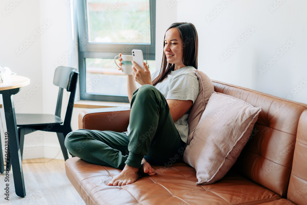 Happy smiling young woman is sitting on the sofa and using the phone.