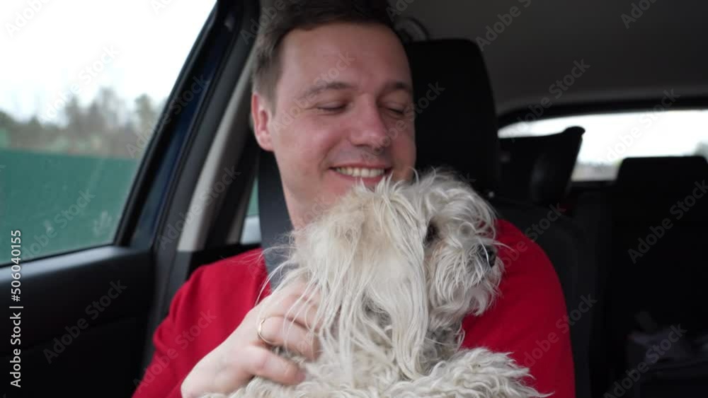 Happy man hugs and strokes his white lapdog, smiles and looking forward. Owner of a fluffy Chinese crested dog travels by car. Travel with pet concept. Happy man hugs and strokes his white lapdog, smiles and looking forward. Owner of a fluffy Chinese crested dog travels by car. Travel with pet concept.