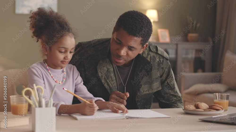 Waist up slowmo of Black man in military uniform helping little daughter with homework, sitting together at desk in living room Waist up slowmo of Black man in military uniform helping little daughter with homework, sitting together at desk in living room