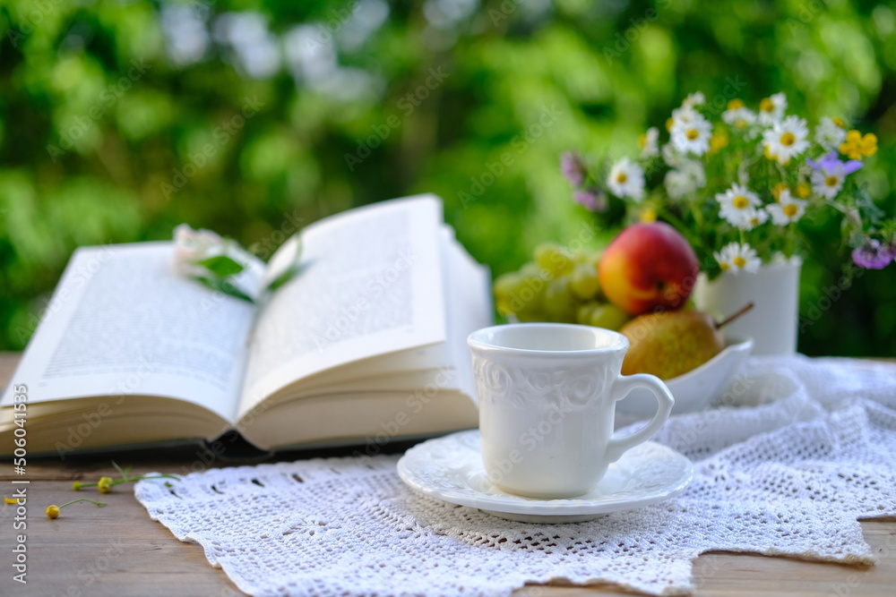 tea, coffee in white mug on saucer, open book, fruits, lace napkin, bouquet of wild flowers on wooden table in garden, blurred natural background with green foliage, concept tea time