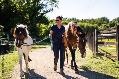 Truly happy smiling farmer woman leading two horses toward camera on beautiful sunny spring day. Countryside horse ranch life.