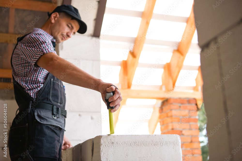 Construction worker at construction site measures the length of window opening and brick wall with tape measure. Cottage are made of porous concrete blocks, work clothes - jumpsuit and baseball cap Construction worker at construction site measures the length of window opening and brick wall with tape measure. Cottage are made of porous concrete blocks, work clothes - jumpsuit and baseball cap