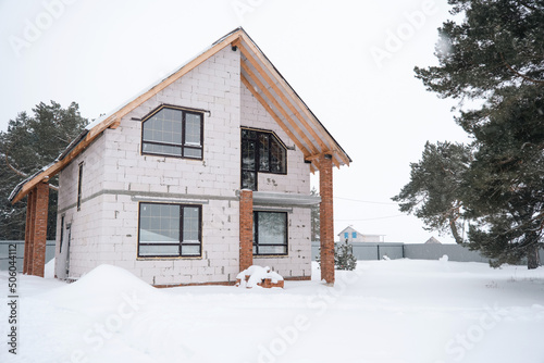 Unfinished construction - a house made of porous blocks in winter, suspension during the construction season, frozen. Snow and facade of the house without exterior decoration with trapezoidal windows