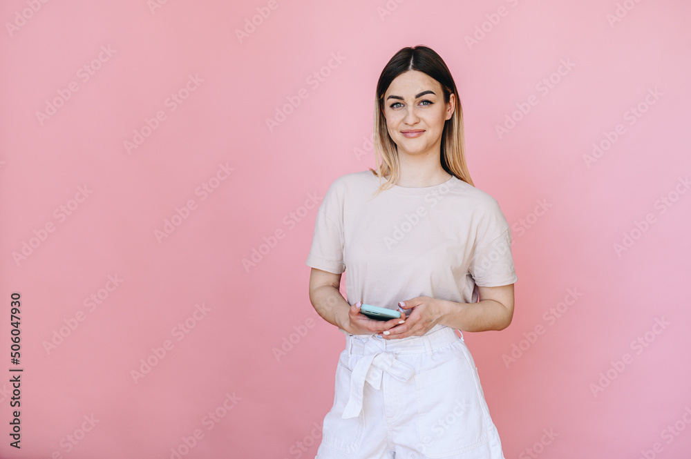 The girl smiles sweetly and stands on a pink background with a phone in her hands in a light T-shirt The girl smiles sweetly and stands on a pink background with a phone in her hands in a light T-shirt