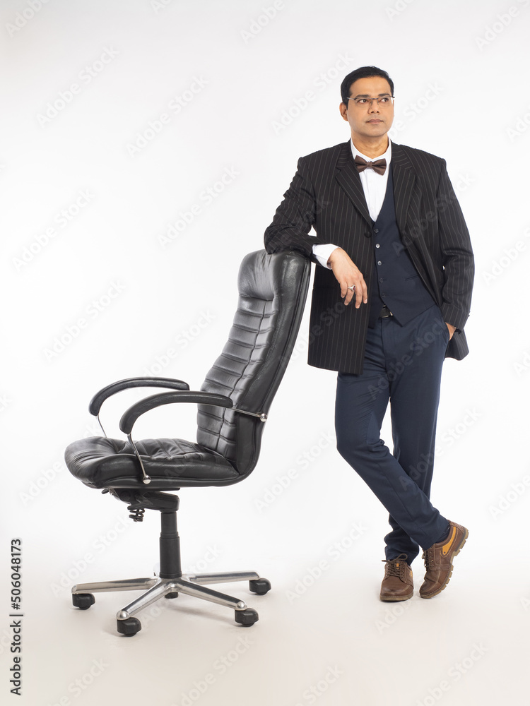 Boss chair. Businessman is standing next to armchair. Man in business suit. Male manager leans on boss chair. Man in suit posing on white background. Concept of selling office furniture. Boss chair. Businessman is standing next to armchair. Man in business suit. Male manager leans on boss chair. Man in suit posing on white background. Concept of selling office furniture.