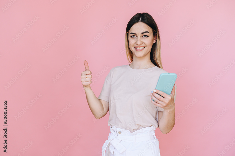 happy girl showing thumbs up and holding a phone in her hand on a pink background happy girl showing thumbs up and holding a phone in her hand on a pink background