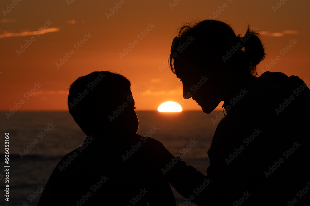 Mother and son at sunset looking at each other tenderly in front of the sea with the sun setting in the background and them silhouetted