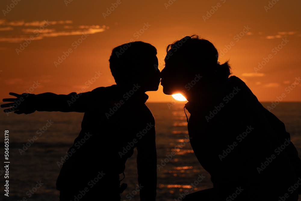 Mother and son at sunset kissing tenderly in front of the sea with the sun setting in the background and them silhouetted