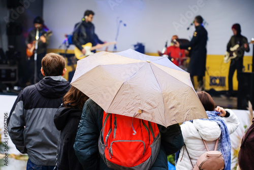 Musical concert in the open area. People with umbrellas stand near the stage. Unrecognizable person. Back view