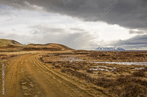 Dirt road in a desolate landscape near lake Myvatn in Iceland with puddles and low vegetation, some hills and a mountain, under a dramatic sky