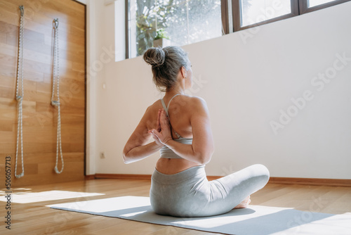View from back on senior female in top stretching arms, keeping hands behind back, doing yoga exercises. Healthy lady sits on fitness mat at home