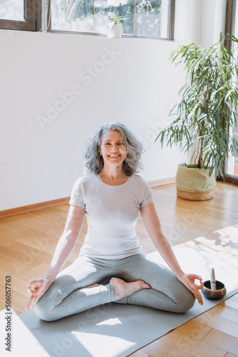 Fototapeta Naklejka Na Ścianę i Meble -  Mindful senior woman sitting cross-legged at the yoga mat, practicing yoga and enjoying meditation. Mature lady with crossed legs meditating at home