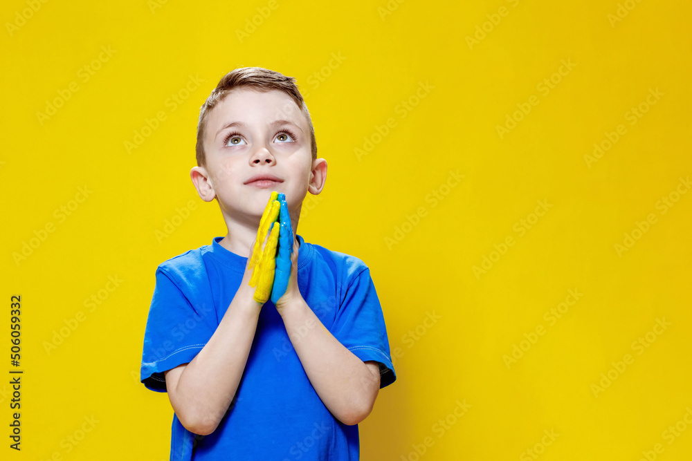 Ukrainian boy prays for Ukraine. Children against war. A boy in a blue T-shirt on a yellow background folded his hands painted in the Ukrainian flag for prayer Ukrainian boy prays for Ukraine. Children against war. A boy in a blue T-shirt on a yellow background folded his hands painted in the Ukrainian flag for prayer