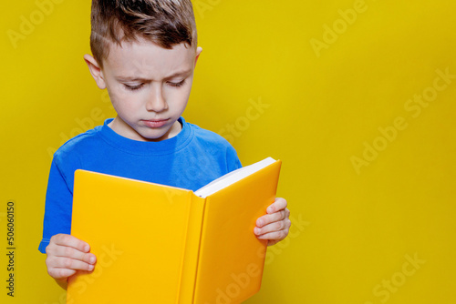 Little cheerful blond green-eyed boy 5-6 years old in a stylish blue T-shirt holding book and reading on yellow wall background, children's studio portrait