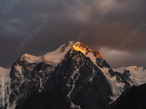 Darkness mountain landscape with great snowy mountain lit by dawn sun among dark clouds. Awesome alpine scenery with high mountain pinnacle at sunset or at sunrise. Big glacier on top in orange light. © sablinstanislav