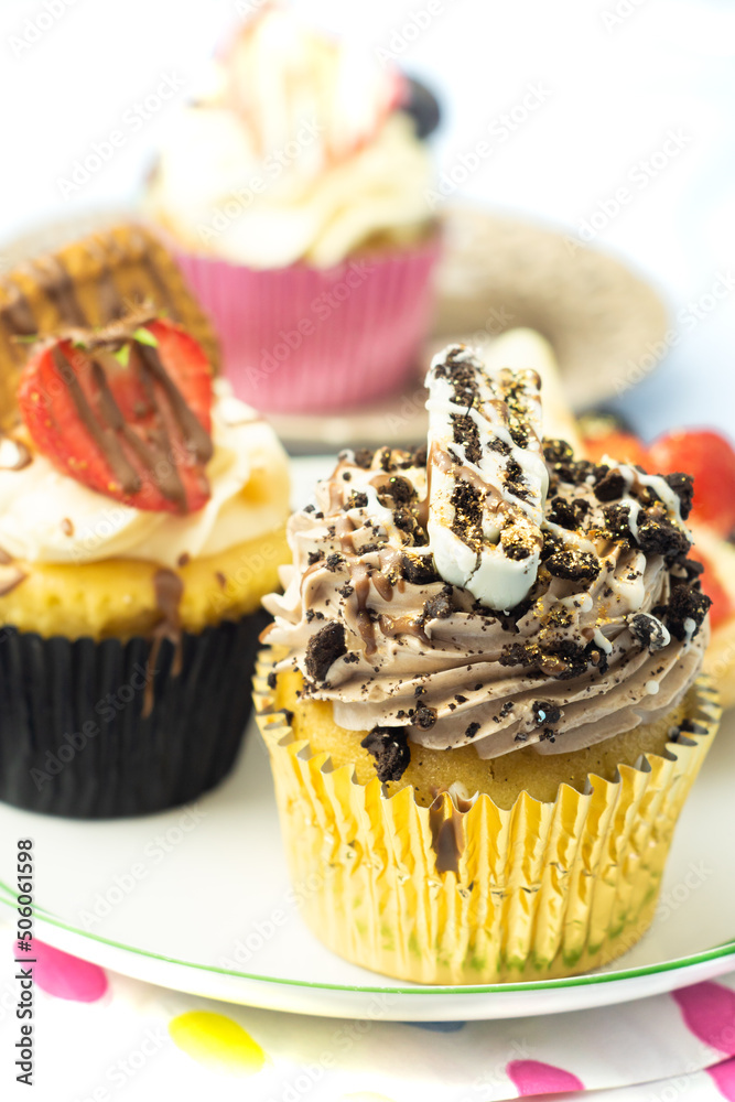 Creamy cupcakes with fruits, cream, oreo cookie and sprinkles in golden wrapping on white plate and light napkin with colorful spots. Nice dessert on pastel background.
