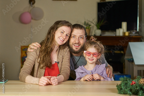 Happy family smiling portrait. A man and a woman hug their daughter. child has fun with his parents at the weekend at home at the table. Hugging is fun. Stylish girl in pink glasses expresses herself.