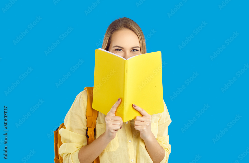 Portrait of happy millennial girl student with backpack on blue studio background hide behind textbook. Smiling young female learner with book enjoy studying course or education. Portrait of happy millennial girl student with backpack on blue studio background hide behind textbook. Smiling young female learner with book enjoy studying course or education.