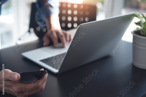 Close up of man using mobile smart phone and working on laptop computer on wooden table at home with blurred background
