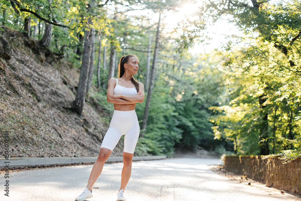 A young beautiful girl in white clothes poses before a running workout, on a road in a dense forest, during sunset. Healthy lifestyle and running in the fresh air. Youth.