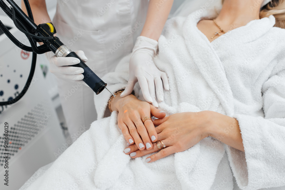 Fototapeta premium A young girl gets carbon peeling for the skin of her hands in a beauty salon. Laser pulses cleanse the skin. Hardware cosmetology. The process of photothermolysis, warming the skin.