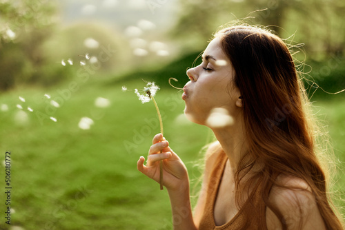 A female freelancer strolls carefree and happily in a green park and blows off a dandelion flower in the sunset light