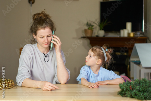 Woman speaks on phone home at table. Serious caucasian woman found out sad news on phone. Work from home with child. girl wants to play with mother. concept working on maternity leave or remote work.