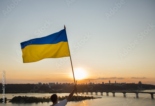 big Ukrainian flag flutters against the backdrop of the sunset sky and cityscape. A man stands on roof of house in Kyiv and holds flag. National symbol of freedom and independence. Stop the war