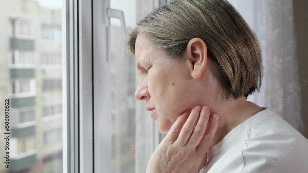 Old lady standing near window touches neck and feels pain. Woman with short haircut looks out of window and palpates painful tumor on neck Old lady standing near window touches neck and feels pain. Woman with short haircut looks out of window and palpates painful tumor on neck